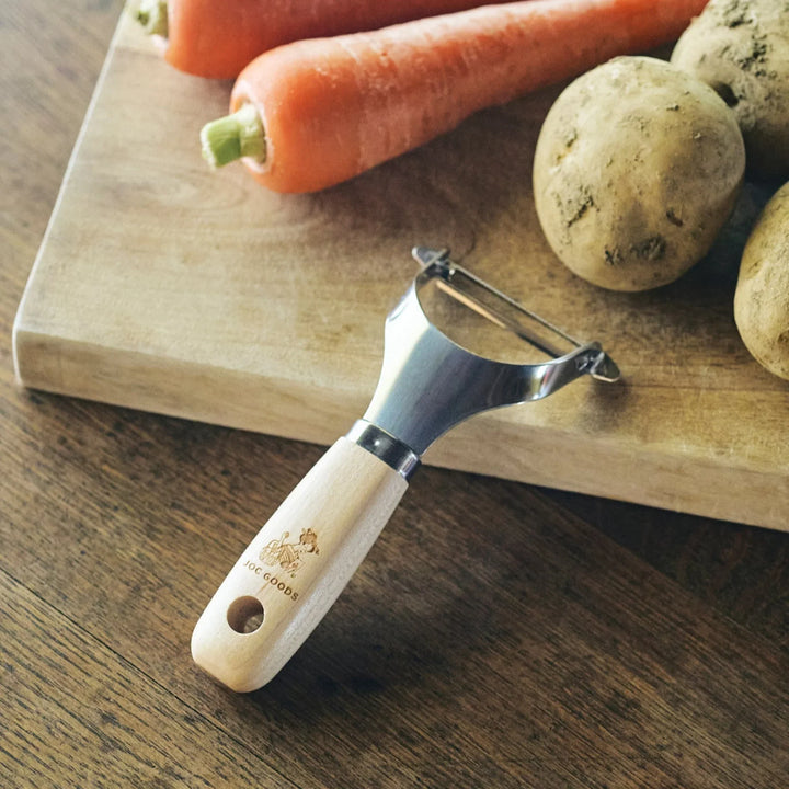 Japanese Wood Handle Peeler with stainless steel blade, placed on a rustic wooden table beside carrots and potatoes on a cutting board.