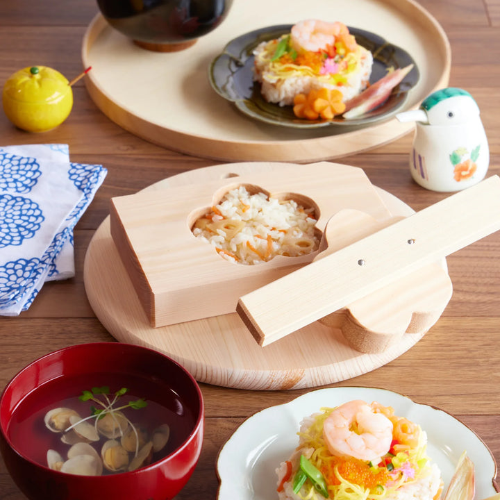 Plum blossom-shaped onigiri mold made of hinoki wood, shown with chirashi sushi and clam soup in a traditional Japanese setting.