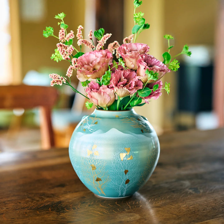 Hand-painted round turquoise vase with gold leaf motifs filled with ruffled pink blooms and green sprigs on wooden tabletop.