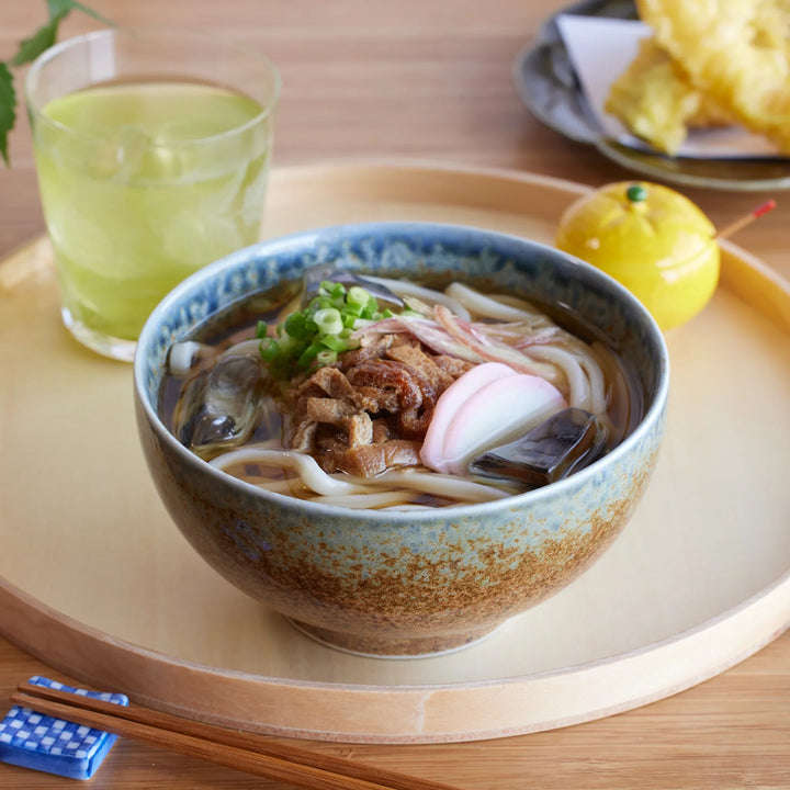 A rustic indigo ceramic donburi bowl filled with udon, garnished with green onions, kamaboko, and vegetables.