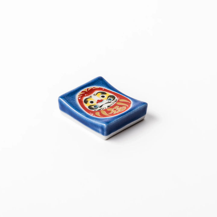 Rectangular Japanese ceramic chopstick rest featuring a red Daruma doll on a blue background, symbolizing perseverance, luck, and good fortune.