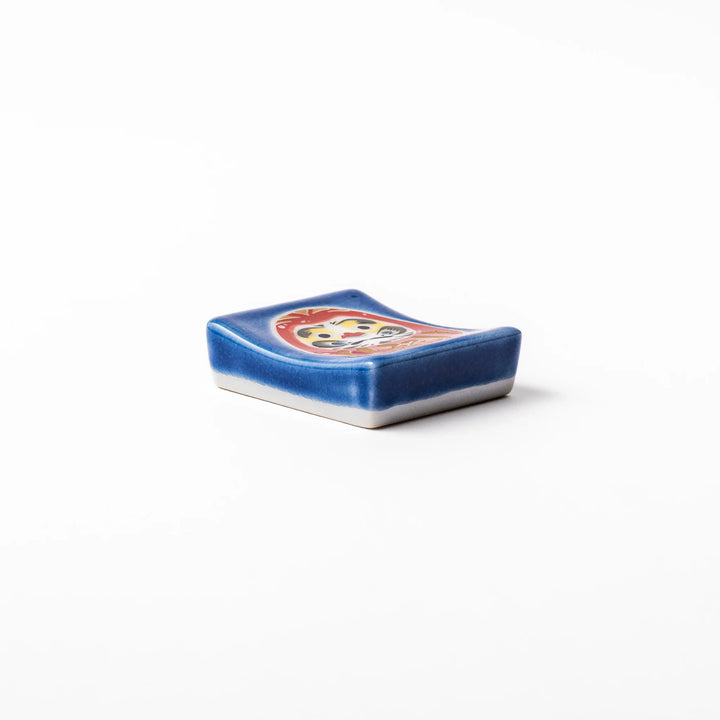 Rectangular Japanese ceramic chopstick rest featuring a red Daruma doll on a blue background, symbolizing perseverance, luck, and good fortune.