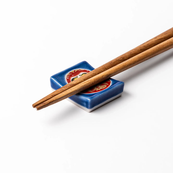 Rectangular Japanese ceramic chopstick rest featuring a red Daruma doll on a blue background, symbolizing perseverance, luck, and good fortune.