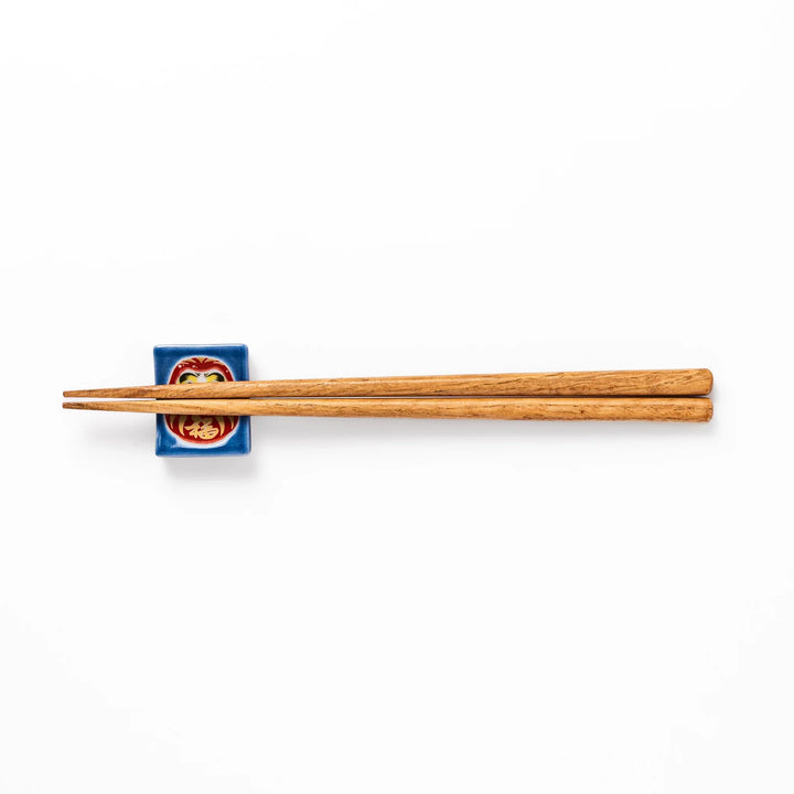 Rectangular Japanese ceramic chopstick rest featuring a red Daruma doll on a blue background, symbolizing perseverance, luck, and good fortune.