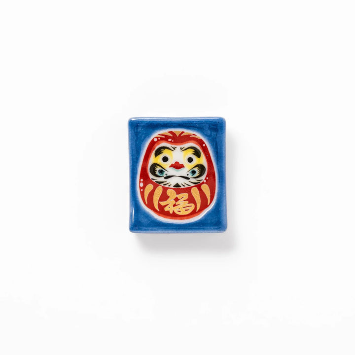Rectangular Japanese ceramic chopstick rest featuring a red Daruma doll on a blue background, symbolizing perseverance, luck, and good fortune.