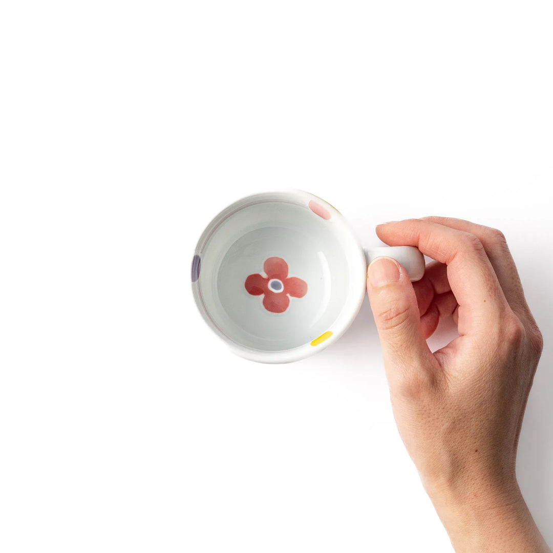 White ceramic kids mug with pink, yellow, red, and purple flower motifs, round handle, shown held for scale