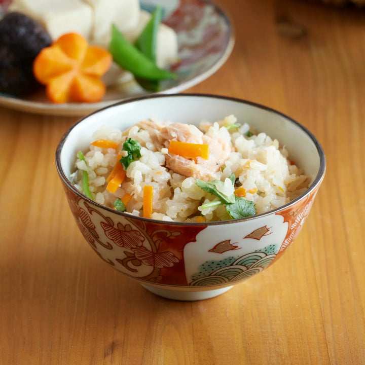 Close-up of a Japanese rice bowl with takikomi gohan, featuring salmon, carrots, and greens in a decorative red-patterned bowl.