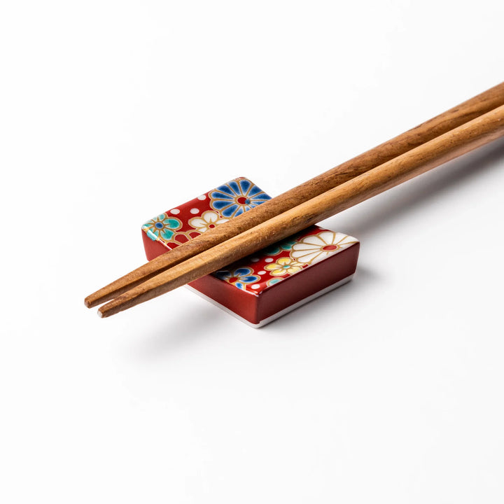 Rectangular Japanese ceramic chopstick rest with a red background decorated with colorful plum and chrysanthemum blossoms, perfect for elegant dining settings.