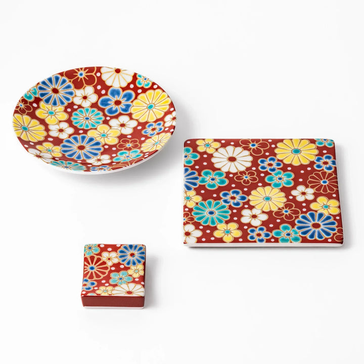 Japanese ceramic tableware set with red background featuring colorful plum and chrysanthemum blossom motifs, including sauce dish, coaster, and chopstick rest.