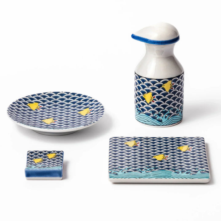 Japanese ceramic tableware set featuring blue wave pattern with yellow shorebirds, including soy sauce dispenser, sauce dish, chopstick rest, and coaster.