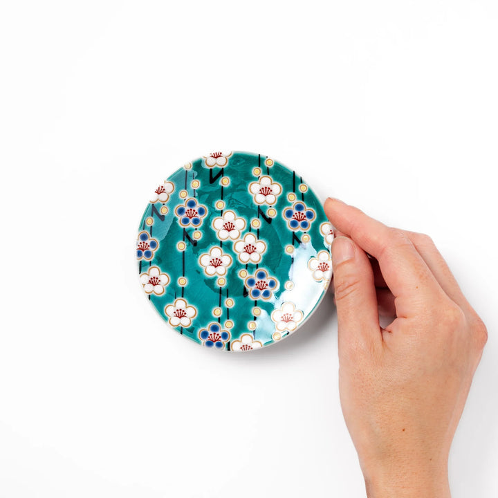 Round Japanese ceramic sauce dish with teal background, decorated with white and blue plum blossom motifs, perfect for serving condiments or small bites.