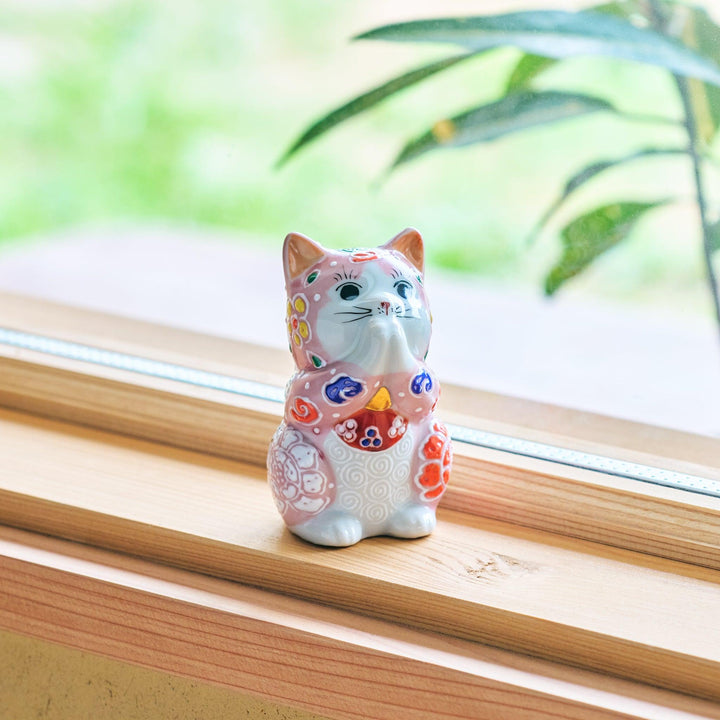 A ceramic maneki-neko (lucky cat) figurine with a pink and multicolored kimono, sitting on a wooden windowsill.