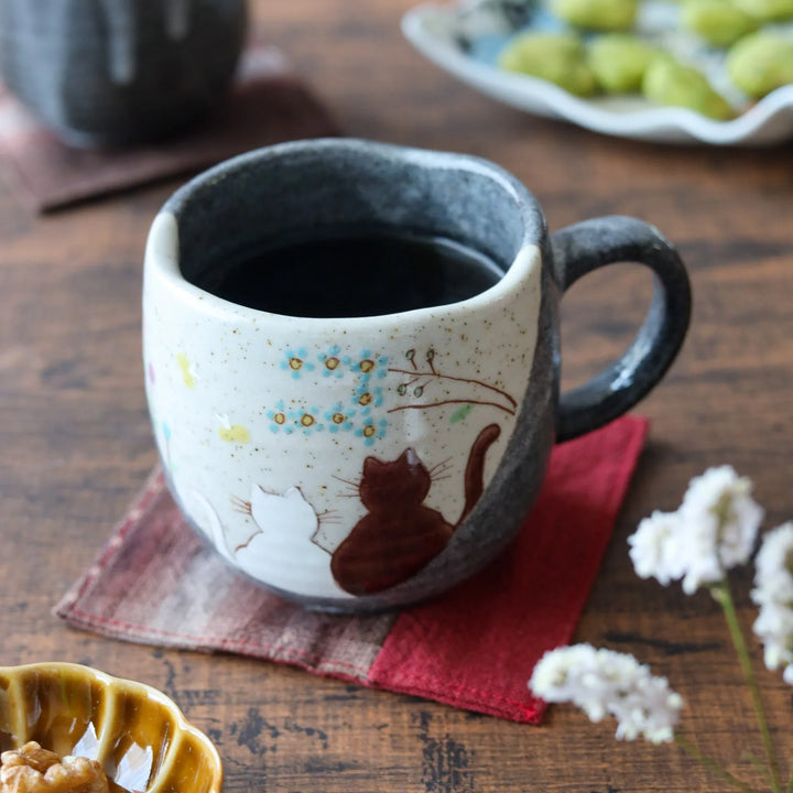 Ceramic mug featuring two cats gazing at blooming flowers under a tree branch, blending soft cream and charcoal tones for a cozy, whimsical touch.