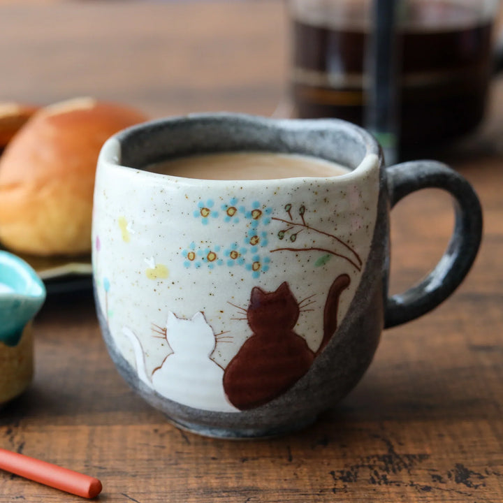 Handcrafted ceramic mug filled with coffee, featuring two cats gazing at blue blossoms, blending playful charm with Japanese artisan detail.