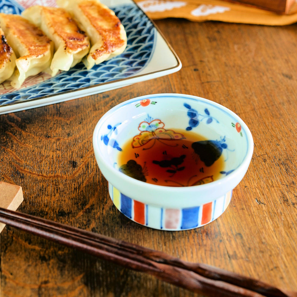 Colorful sauce dish with raised edges, featuring a butterfly and floral pattern inside and vertical red, blue, and yellow stripes outside, filled with dipping sauce.