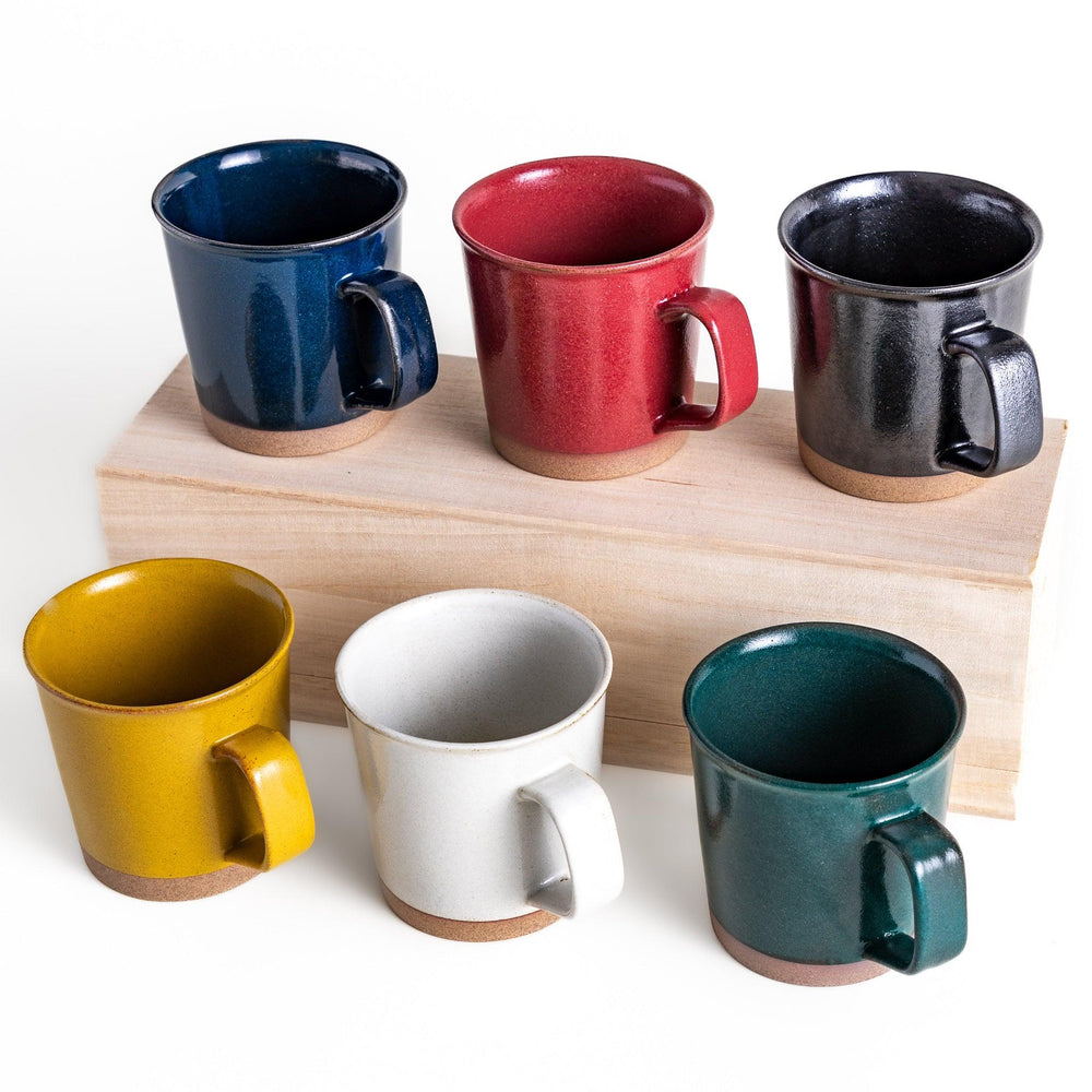 A set of six colorful mugs in blue, red, green, yellow, white, and black, each with a smooth, glossy finish and a natural clay base, arranged neatly on a wooden block.