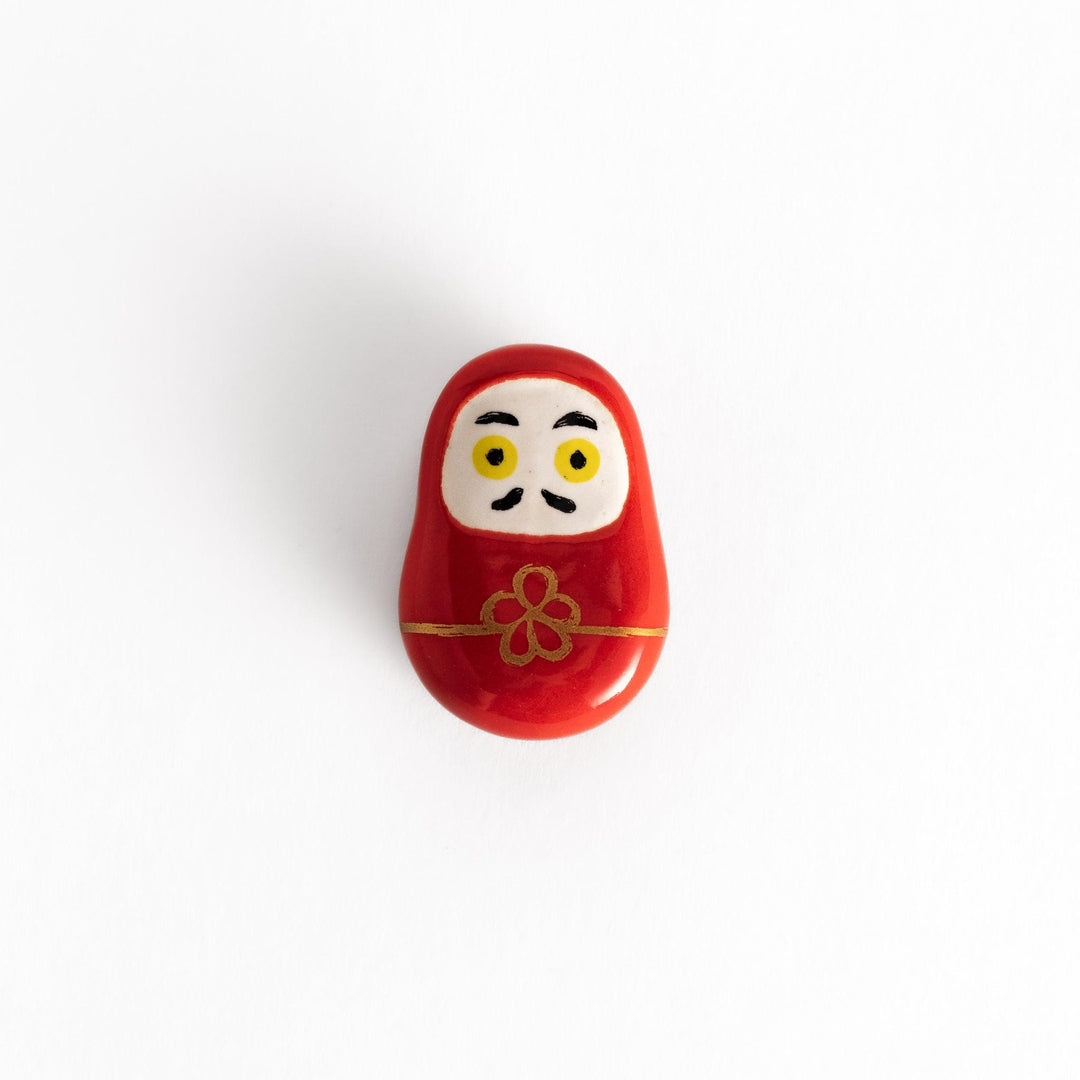 A red daruma doll-shaped chopstick rest with a decorative gold cord and flower design, featuring bold yellow eyes on a white face.