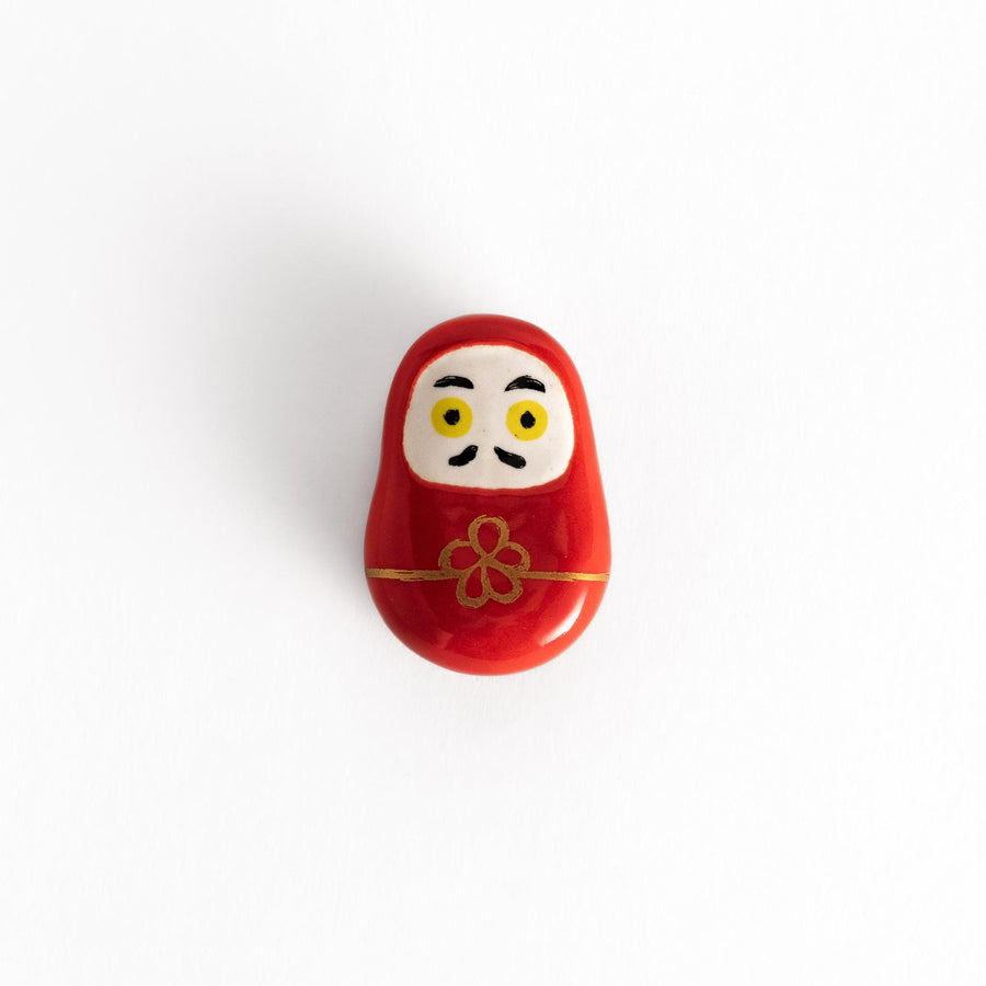 A red daruma doll-shaped chopstick rest with a decorative gold cord and flower design, featuring bold yellow eyes on a white face.