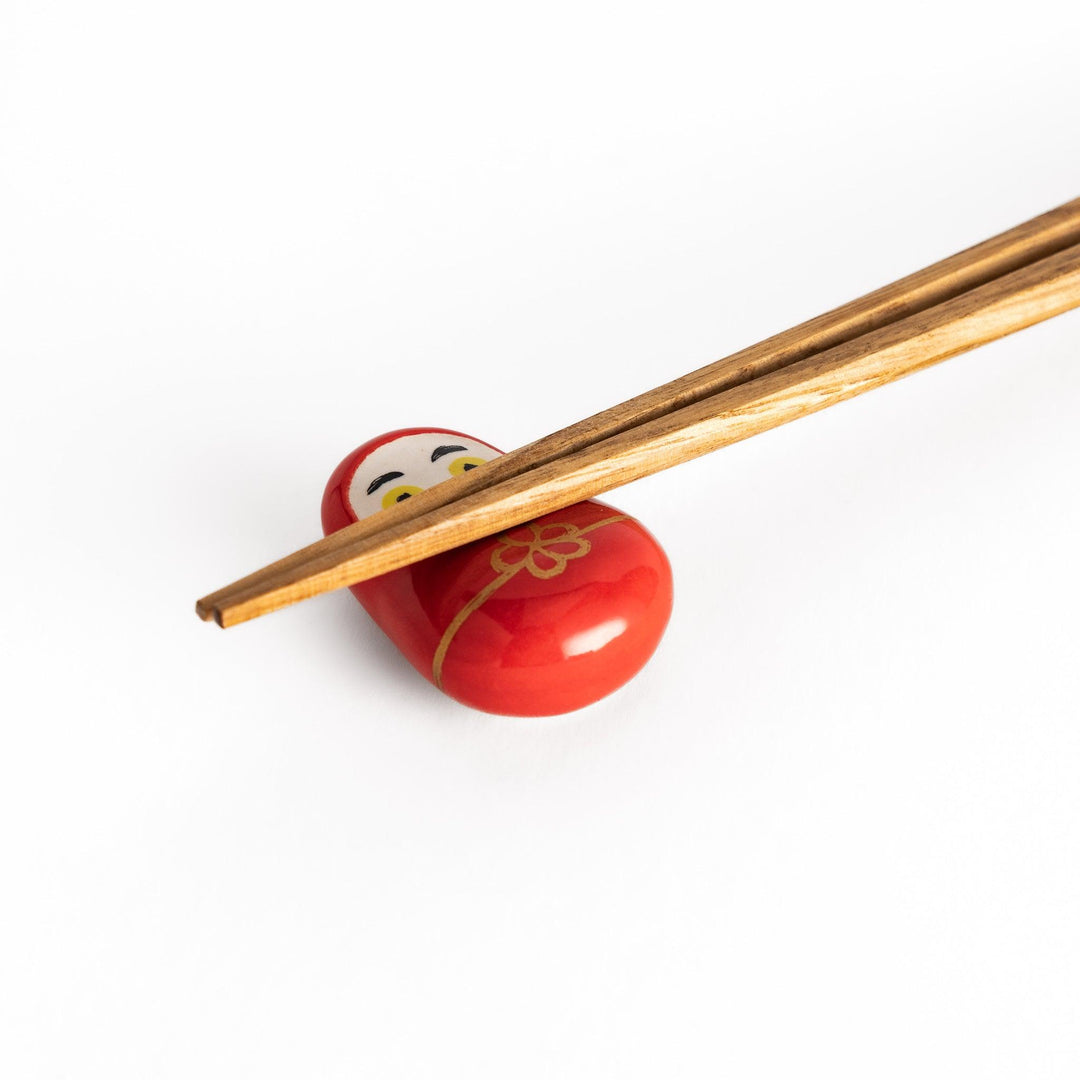 A close-up view of chopsticks resting on the red daruma chopstick rest, highlighting the gold flower design and glossy finish.
