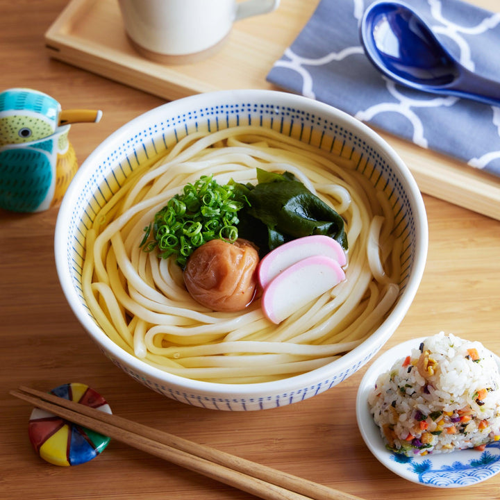 Udon noodle soup in a blue patterned bowl, topped with green onions, wakame, umeboshi, and sliced kamaboko in a clear broth.