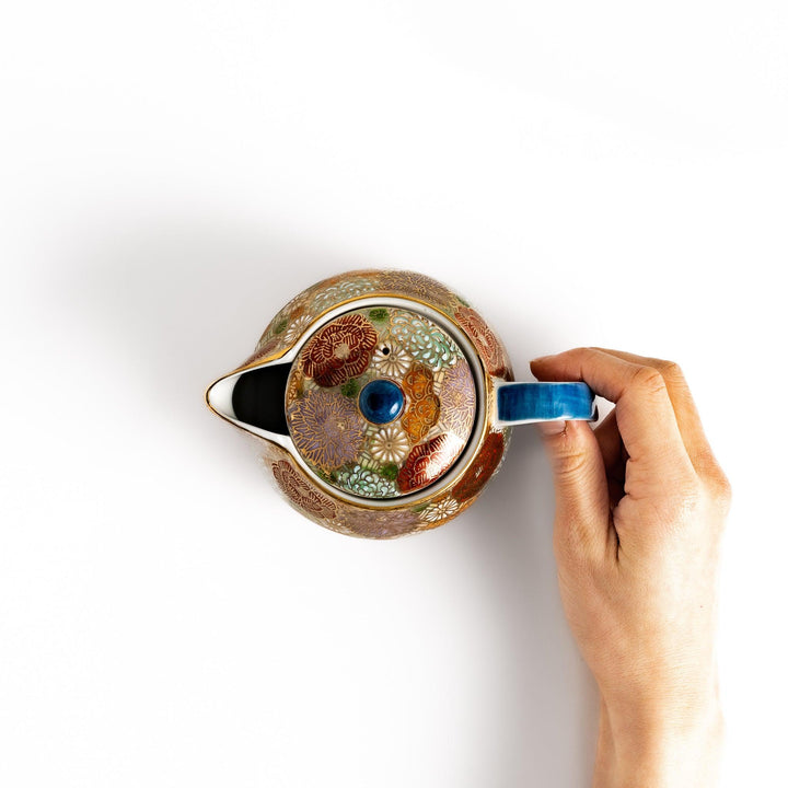 Hand holding the handle of a ceramic teapot with a vibrant floral pattern in gold and pastel colors, viewed from above. The teapot features a blue lid.