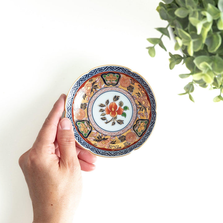 A round sauce dish with a central red floral motif, surrounded by a detailed border of gold, blue, and green floral patterns.