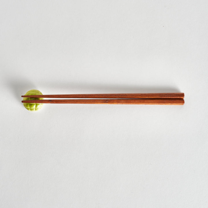 A pair of wooden chopsticks gently picking up a green melon-shaped chopstick rest with a textured melon rind design.