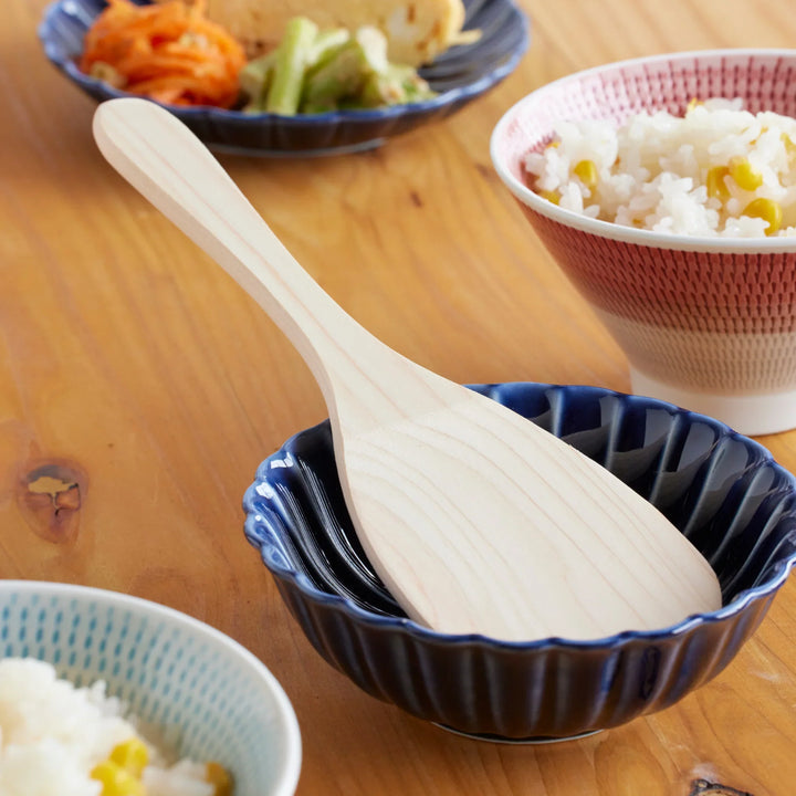 Japanese hinoki wood rice paddle placed in a blue dish, shown with freshly cooked rice and side dishes for authentic everyday dining.