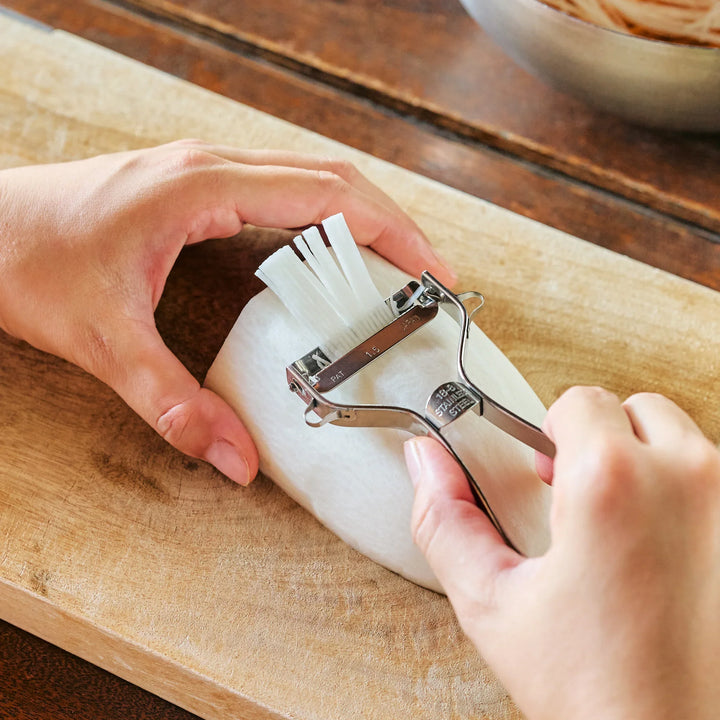 A hand using a Japanese julienne peeler to shred daikon radish into fine strips (Tsuma) on a wooden board.