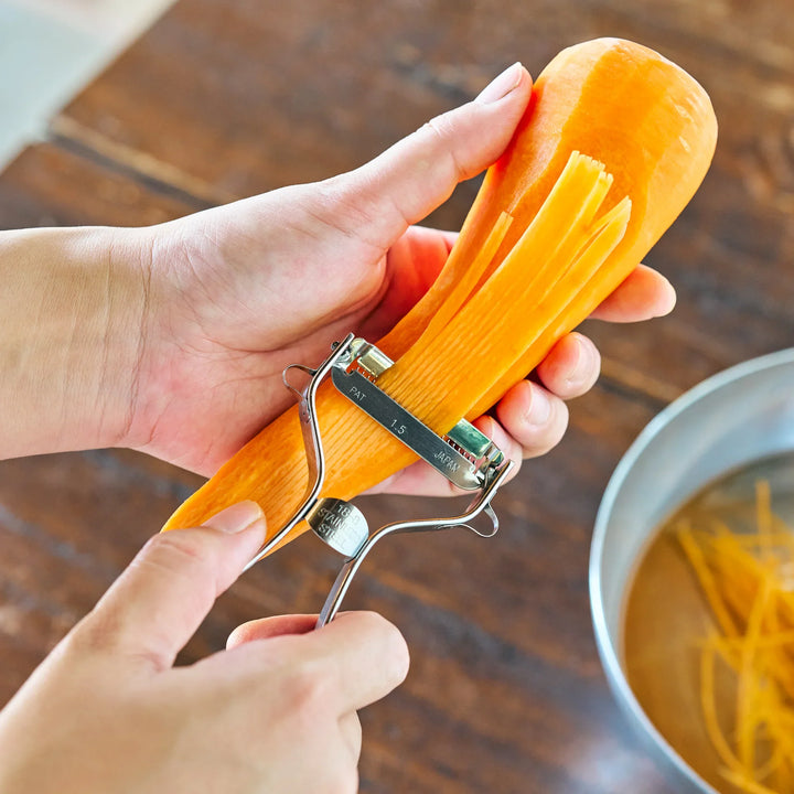 A person shredding a carrot into thin strips with a Japanese julienne peeler over a metal bowl.