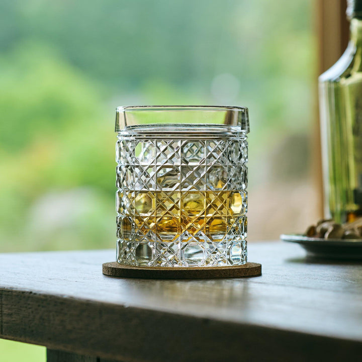 Cut Japanese whiskey glass with a lattice pattern, filled with whisky and ice, on a cork coaster near a window.