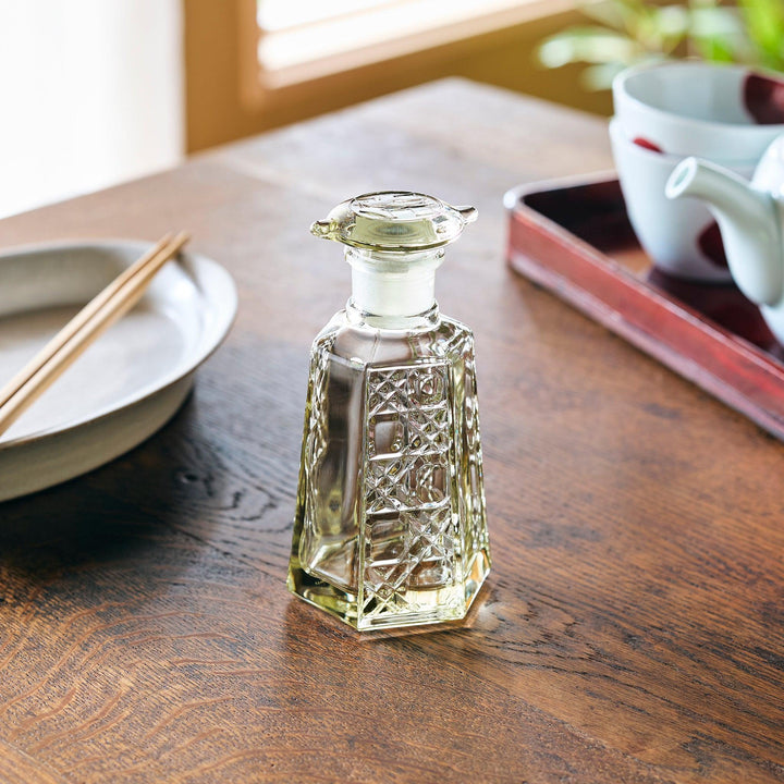 A hexagonal glass soy sauce dispenser with intricate lattice cuts, filled with light-colored liquid on a wooden table.