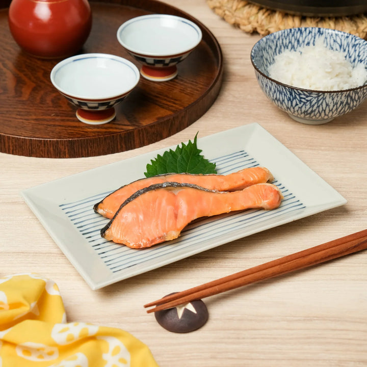 Grilled salmon fillets on a rectangular Japanese plate, served with rice, chopsticks, and sake cups for a traditional meal.