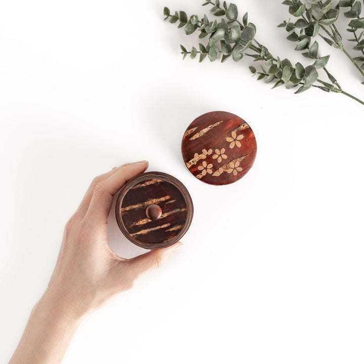 Hand holding a cherry bark tea canister with a lid decorated with cherry blossoms, a beautiful and functional container for storing tea leaves.