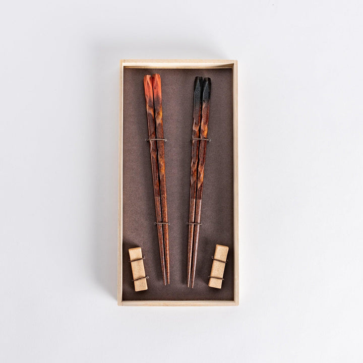 Boxed set of dark brown chopsticks with textured designs and matching rests, placed neatly in a wooden box on a dark background.