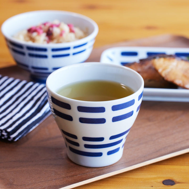 Ceramic cup with indigo horizontal patterns, filled with green tea, placed on a wooden tray with matching tableware