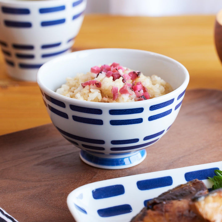 A ceramic rice bowl with indigo horizontal patterns, filled with rice topped with pink pickles, on a wooden tray.