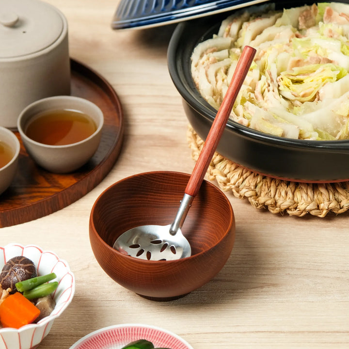 Perforated stainless steel soup ladle with a wooden handle resting in a wooden bowl, placed beside a navy Japanese clay pot filled with layered hot pot for traditional dining.