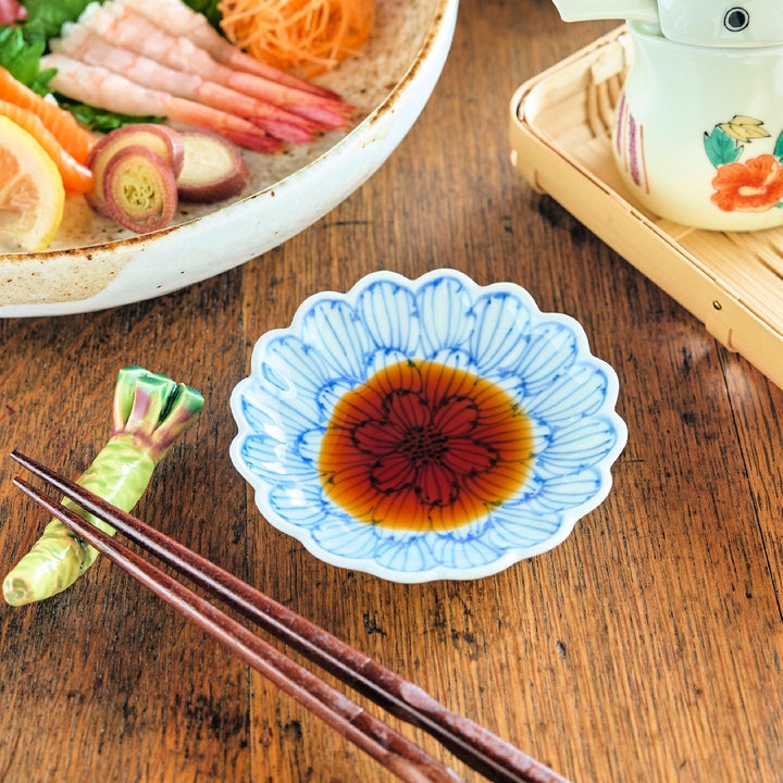 Scalloped edge sauce dish with a vivid red and blue petal pattern, filled with soy sauce and placed on a wooden table beside chopsticks and wasabi chopstick rest.