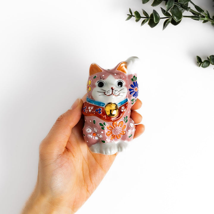 Charming pink blossom Maneki Neko figurine, adorned with colorful floral patterns and a golden bell, symbolizing good luck and prosperity with a cheerful waving gesture.