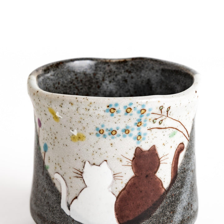 Charming ceramic teacup featuring two cats gazing at small flowers, with a unique speckled glaze and a comfortable shape for holding.