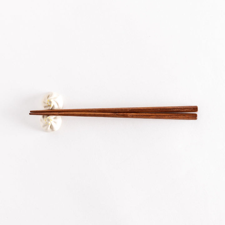 Wide shot of wooden chopsticks resting on a soup dumpling-shaped chopstick rest, placed on a white surface.