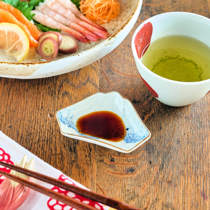 Mount Fuji-shaped mini sauce dish with blue sprayed ink pattern, filled with soy sauce and placed beside a teacup and sashimi on a wooden table.