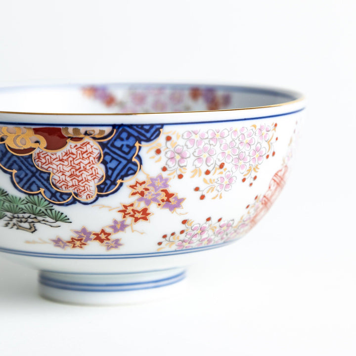 A round ceramic rice bowl with detailed traditional Japanese designs of trees and clouds in blue, red, and green on a white background.
