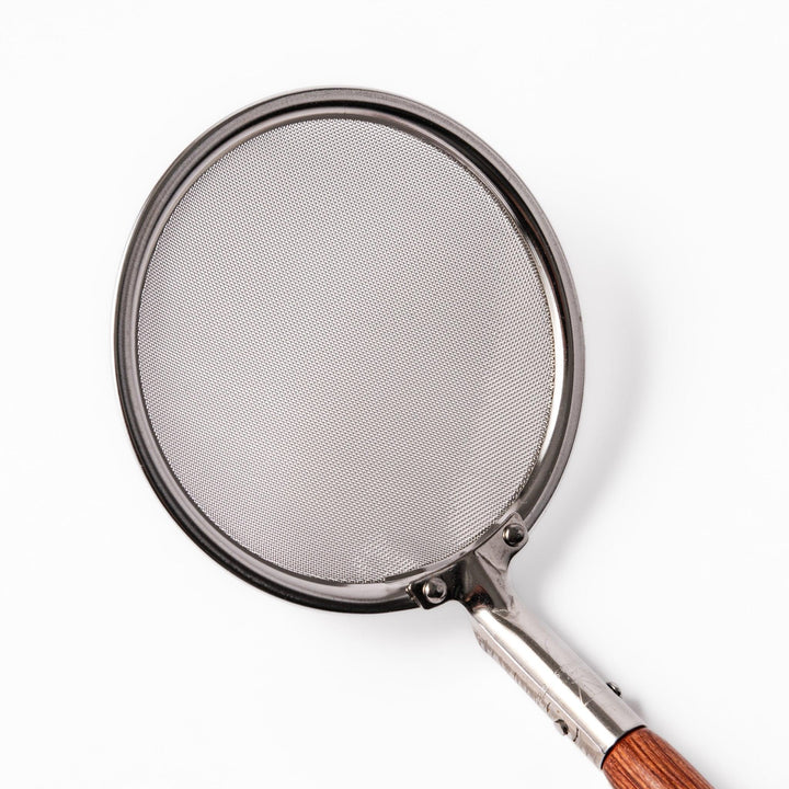 Stainless steel round-mesh skimmer with long wooden handle, perfect for straining soups and frying in home kitchens.