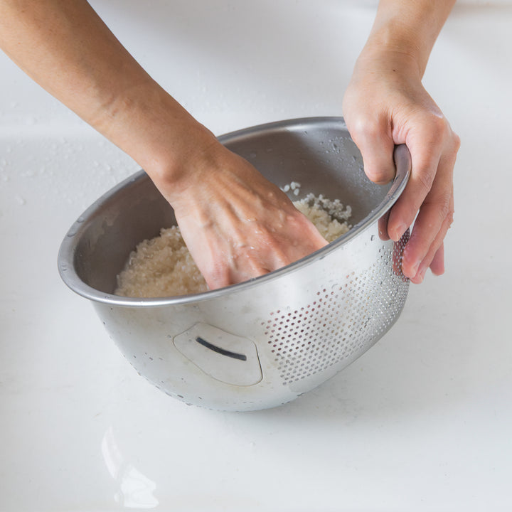 Nami washing rice by hand in a stainless steel colander, swirling the grains to clean them evenly.