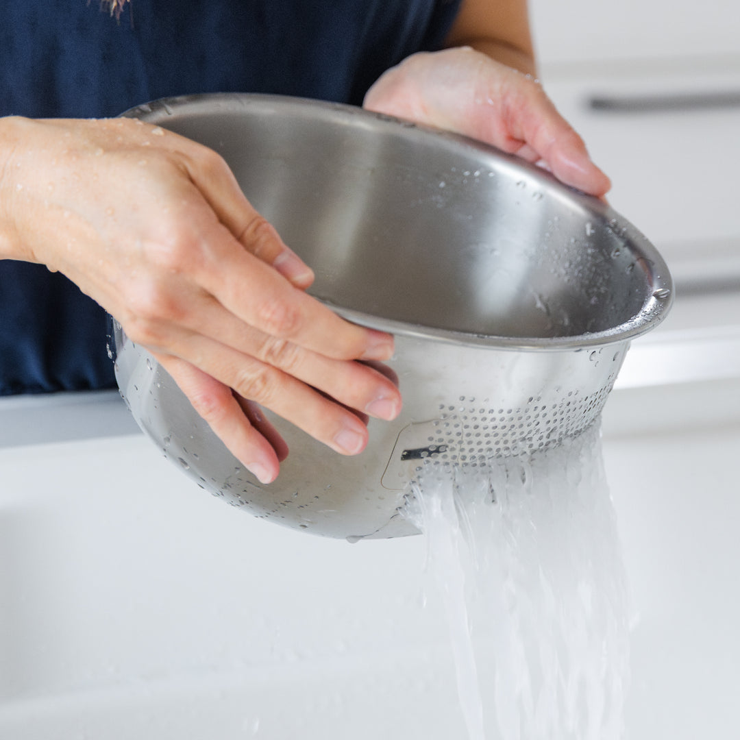 Nami draining cloudy water from freshly washed rice using a stainless steel colander in the sink.