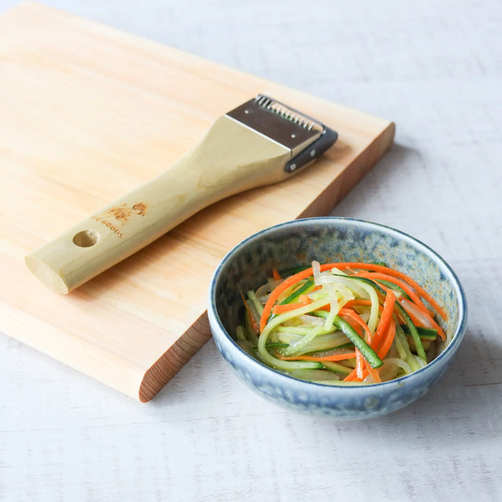 A wood handle julienne peeler on a cutting board beside a bowl of thinly sliced cucumber, carrot, and onion salad.