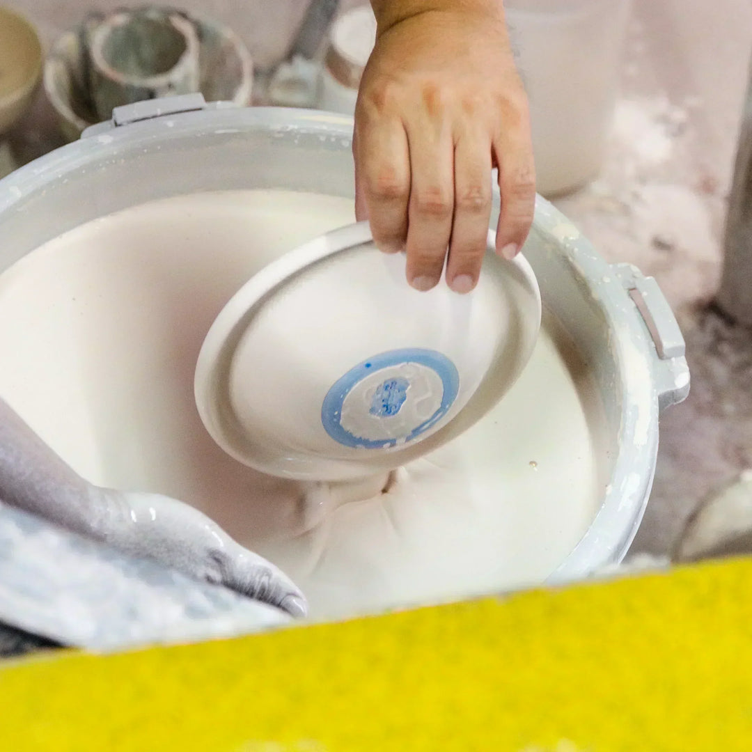A craftsman dipping a ceramic bowl into a creamy white glaze, showing the glazing process with hands steady over a large vat