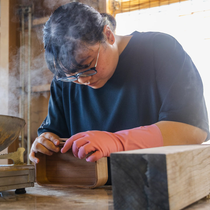 A craftsperson shaping a wooden box with gloved hands in a workshop filled with steam, focused closely on the fine details